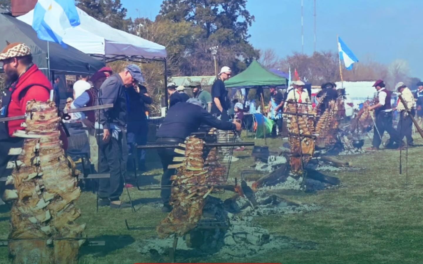 La imagen muestra a varios asadores al aire libre cocinando costillares y cortes de carne ensartados en estacas de hierro, dispuestas en círculo alrededor de fogatas sobre el suelo. En el fondo se aprecian banderas argentinas, carpas y personas circulando en un predio verde. En la parte inferior, sobre un fondo verde translúcido, se lee en letras blancas: "PARA AGENDAR / PUEBLO ANDINO SE PREPARA PARA LA 3RA EDICIÓN DE VACÍO A LA ESTACA".