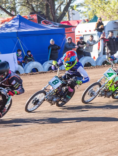 Tres motociclistas de speedway compiten en una pista de tierra durante el día. El piloto central viste traje azul con casco multicolor e inclina su moto con el número 8 en una curva pronunciada, rodeado de público y carpas de fondo.