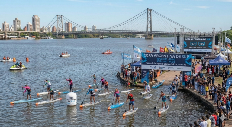 Esta imagen muestra un grupo de personas practicando paddle surf (SUP) con el Puente Colgante de Santa Fe al fondo.