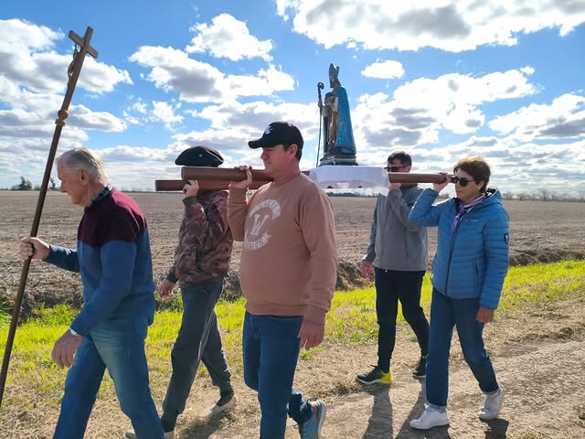 Un grupo de personas avanza en una procesión religiosa a pie por un camino de tierra rural bajo un cielo parcialmente nublado.En el centro, cuatro personas cargan sobre sus hombros una tarima de madera que sostiene una estatua de un santo con vestiduras azules y mitra episcopal. Al frente del grupo, un hombre mayor camina liderando la marcha mientras sostiene en su mano una cruz procesional de madera.