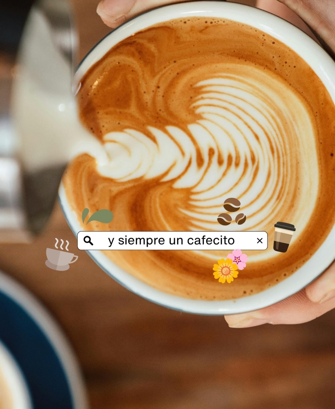 Vista aérea en primer plano de una taza de café donde se está vertiendo leche para crear un diseño de arte latte (rosetta). Sobre la espuma sobresale una barra de búsqueda estilizada que dice "y siempre un cafecito", decorada con stickers e iconos ilustrados de granos de café, flores y tazas humeantes.