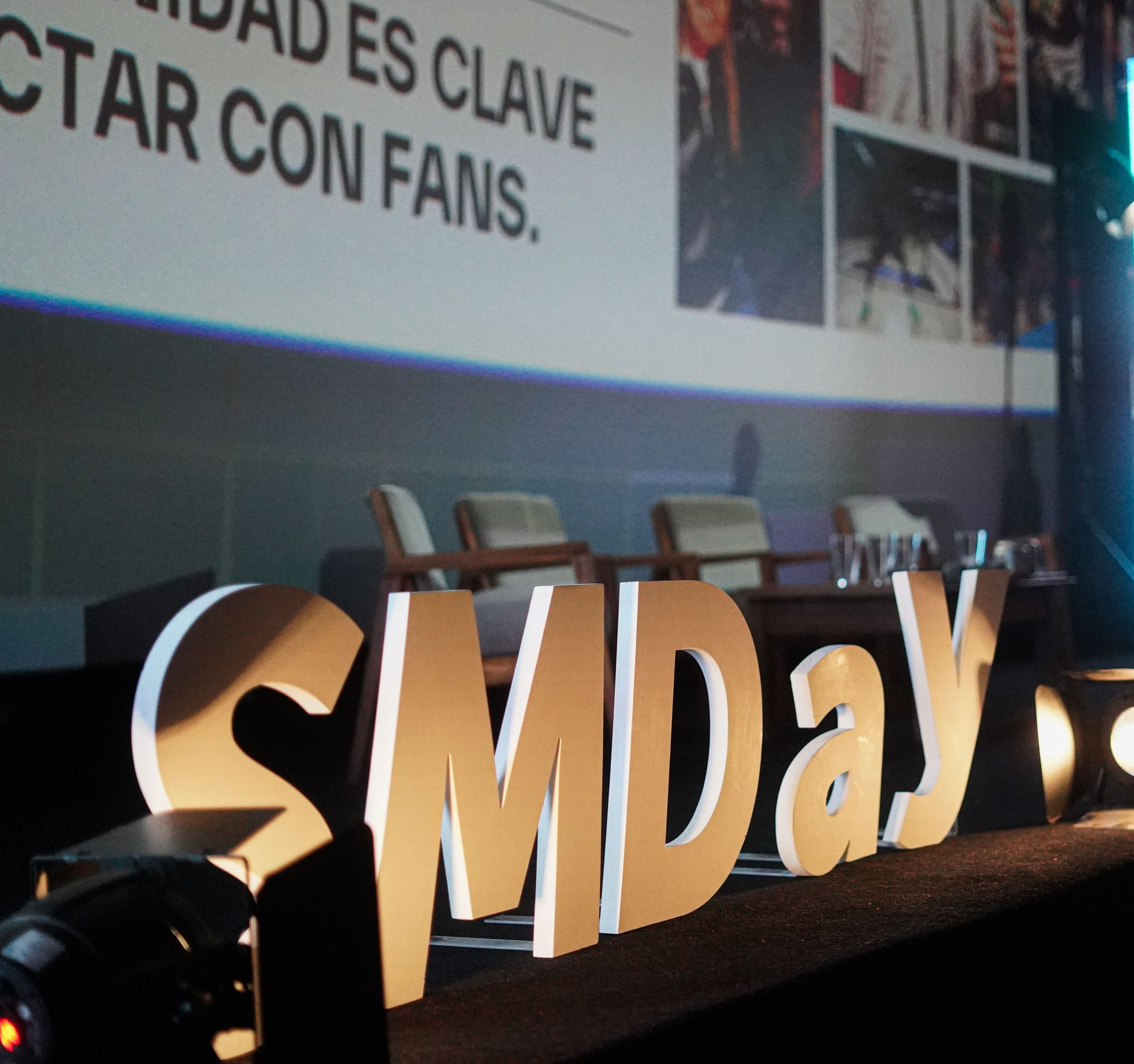 Un cartel corpóreo con las letras "SMDay" iluminadas se destaca en el borde de un escenario preparado para un evento de comunicación digital.De fondo se observa una gran pantalla de proyección que muestra una presentación con el texto en mayúsculas "...DAD ES CLAVE" y "CTAR CON FANS.", junto con imágenes de creadores de contenido. En la tarima, detrás de las letras corpóreas, se encuentra dispuesto un panel de conversación vacío con varias sillas y una mesa larga para los disertantes, ambientado con una iluminación profesional de tonos azulados.