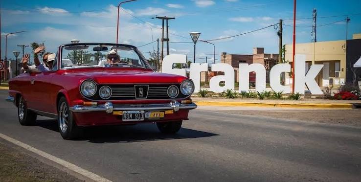 Fotografía a plena luz del día de un automóvil clásico e histórico de marca IKA Torino, modelo descapotable o convertible, pintado en color rojo brillante. El vehículo transita sobre una calle de pavimento con sus luces delanteras redondas encendidas. En el interior se encuentran tres personas: el conductor lleva puesto un sombrero claro y gafas de sol, mientras que los pasajeros del asiento trasero saludan con el brazo levantado hacia la cámara.