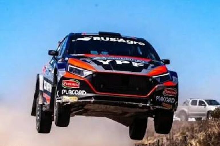 Un auto de rally Hyundai i20 salta por el aire durante una carrera del Rally Argentino. El vehículo, decorado con los logos de YPF y RUS Agro, vuela sobre un camino de tierra con las sierras y el cielo azul de fondo.